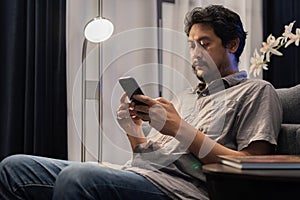 Shot view of man reads online book on smart phone and sitting on soft couch against bright lamp