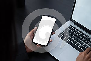 Shot of woman holding blank screen mock up mobile phone during typing on laptop computer.