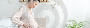 Shot of worried pregnant woman in nightie feeling pain and touching belly while sitting on bed