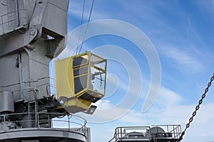 Shot of yellow control cabin with worker. Old, rusty, grey port crane lifting cargo in ship on clear blue sky background.