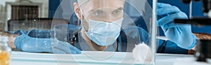 Shot of young biologist in medical mask and latex gloves looking at white mouse in glass box