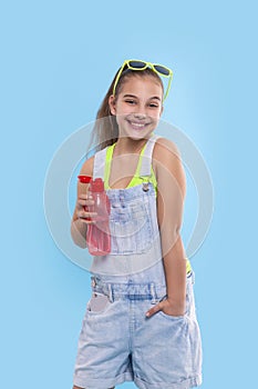 A young brunette girl wearing  denim overalls shorts stands with  a red bottle of water
