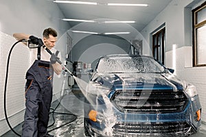 Shot of young Caucasian man worker washing blue car under high pressure water in car wash service. Removing the soap