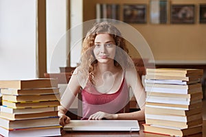 Shot of young foxy haired female student sitting at table in library, preparing for test or exam, posing with pleasant facial