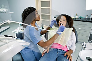 Shot of young pretty female african dentist in blue uniform, making dental check up and caries treatment with drill for