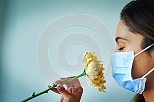 I cant seem to smell anything. Shot of a young woman smelling a flower while wearing a surgical mask.