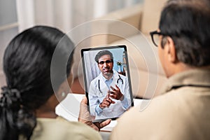 Shoulder shot of Indian Senior couple taking advice from doctor during online video call from tab at home - concept of