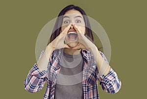 Shouting excited young brunette woman in plaid shirt with hands near mouth on khaki background.