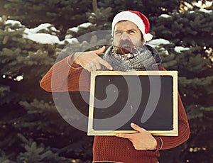 Shouting man in santa claus hat, hipster with beard and moustache covered with white frost, points at blank black board