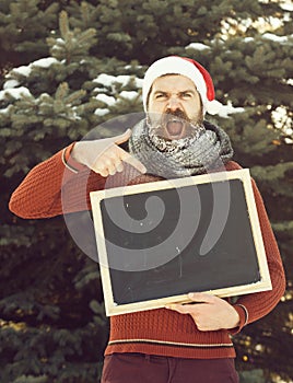 Shouting man in santa claus hat, hipster with beard and moustache covered with white frost, points at blank black board