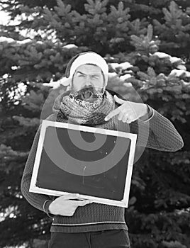 Shouting man in santa claus hat, hipster with beard and moustache covered with white frost, points at blank black board