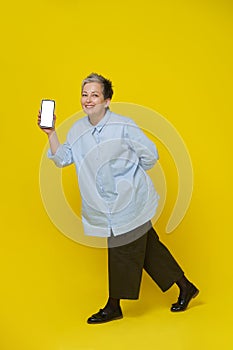 Showing phone in hand mature grey haired woman smiling on camera. Pretty woman in blue shirt and brown trousers isolated