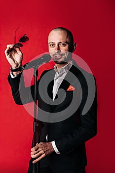 Showman-interviewer with emotions. A young, elegant man holds glasses and a microphone on a red background.The concept