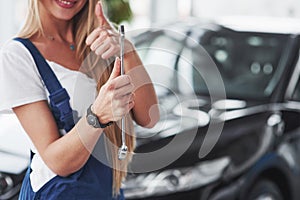 Shows thumb up and holds wrench in other hand. Nice blonde woman repairer is on her work. Indoors at car shop