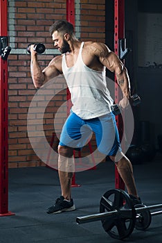 Shredded bodybuilder with a beard is doing biceps curls with dumbbells in a gym