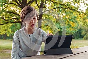 Shrewd boy in light gray longsleeve sitting at wooden table in park and writing task, homework on tablet using stylus.