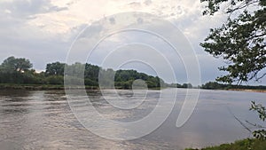 Shrubs, birches and willows grow on the grassy banks of the river. Further away is the forest. A strong wind creates ripples on th