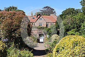 Shrubs and trees either side of a gravel path lead up to an open gateway and onwards to a large house. The sky is blue and