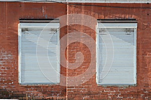 Shutters. Old facade painted red brick wall with 2 windows closed by white roller shutters, illuminated by bright sunlight