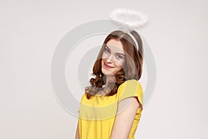 Shy attractive teenager girl with wavy hair in casual yellow T-shirt, standing with halo on her head and looking at camera and