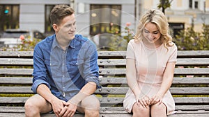 Shy blonde girl smiling, attractive guy flirting with beautiful woman on bench