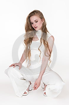 Shy blondie woman sitting squatting position and looking down, wearing white jumpsuit and slingbacks photo