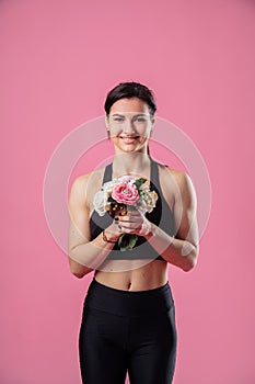 Shy, cute, beautiful, sexy girl fitness trainer is standing in the studio, isolated on a pink plain background with a bouquet of