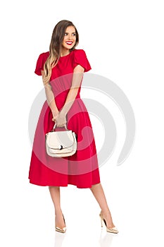 Shy Fashionable Woman In Red Dress, Gold High Heels And Beige Purse Is Looking Away And Smiling