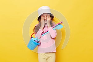Shy funny little cute girl in panama holding beach sandbox toys rake and shovel isolated over yellow background covering eyes with