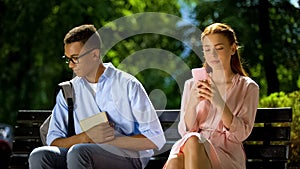 Shy geek sitting on bench with beautiful girl using smartphone, hesitation