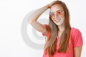 Shy and tender charming redhead lady with freckles in trendy sunglasses and pink blouse combing hair behin with hand on