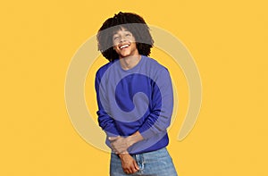 Shy young black guy in blue sweatshirt posing over yellow studio background