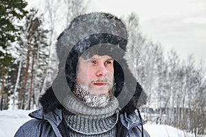 Siberian Russian man with a beard in hoarfrost in freezing cold in the winter freezes and wears a hat with a earflap