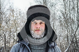 Siberian Russian man with a beard in hoarfrost in freezing cold in the winter freezes and wears a hat with a earflap