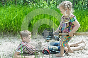 Sibling girl and boy digging on beach dune and burying each other in white sand at pinewood background