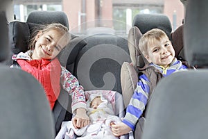 Sibling sitting in car seats and holding doll also in car seat by hand, child transportation safety