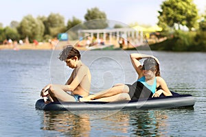 Siblings brother and sister with inflatable matrass swim in the lake on sand beach background