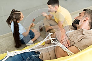 siblings eating chocolate and father lying tied on sofa at home