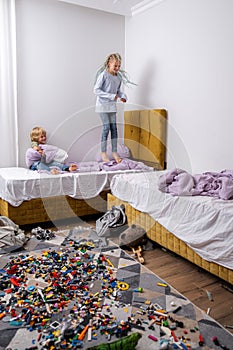 siblings having fun, laughing, boy and girl jumping on bed in messy childrens room, kids playing among the many toys at