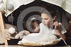 Siblings lie in hut of chairs and blankets. Brother and sister reading book with a flashlight at home