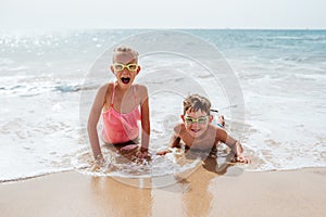 Siblings playing on beach, lying in water, having fun. Smilling girl and boy in swimsuits, swimming googles on sandy