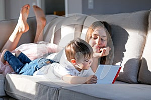 Siblings reading book together on couch with feet up. Hard light. Sister and her little brother spending time together at home rea