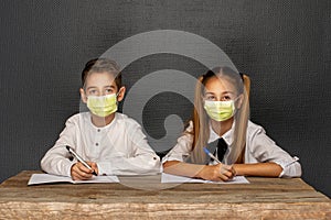 Siblings, schoolchildren are sitting at a school desk over black wall l in medical protective masks. Quarantine at school. Back to