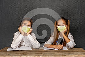 Siblings, schoolchildren are sitting at a school desk over black wall l in medical protective masks. Quarantine at school. Back to