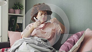 Sick african american girl with flu lying on sofa at home. Ill young black woman with cold checking temperature with