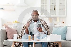 Sick african american man measuring body temperature and texting with doctor on smartphone, having online consultation