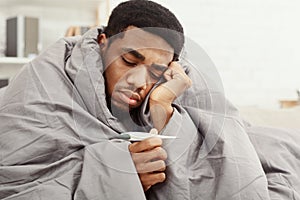 Sick african-american man wrapped in blanket holding thermometer