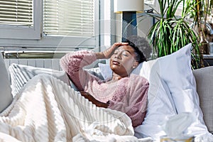 Sick African American woman with headache sitting under the blanket. Sick woman covered with a blanket lying in bed with seasonal