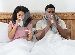 Sick african couple sitting in bed, measuring fever, sneezing noses