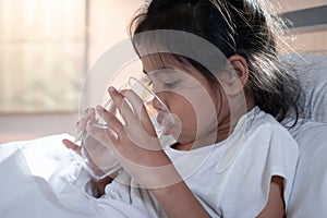 Sick asian child girl is drinking water from a glass after eat medicine in bedroom.